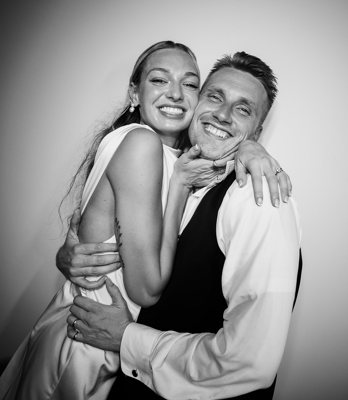 Three people, including one in a wedding dress and another in a suit, pose happily in front of a white backdrop under a chandelier.