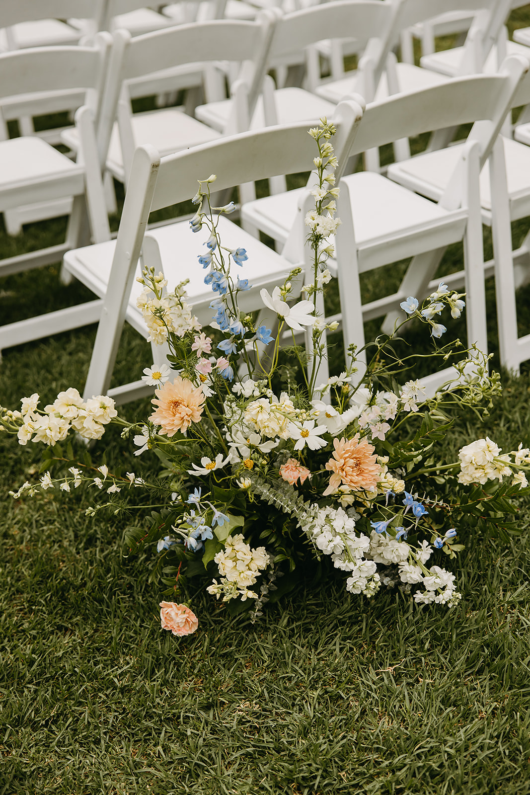 Floral arrangement with pastel flowers on grass near white folding chairs.