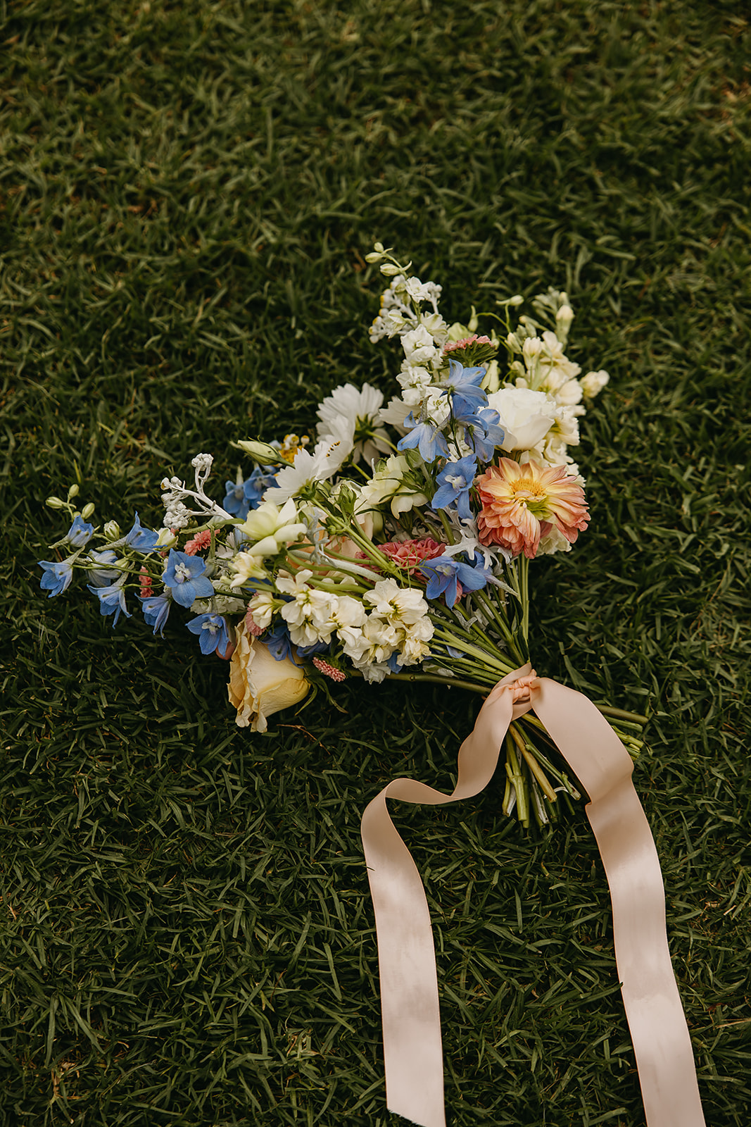 A bouquet of mixed flowers with blue, white, and pink blooms, tied with a ribbon, lies on green grass.