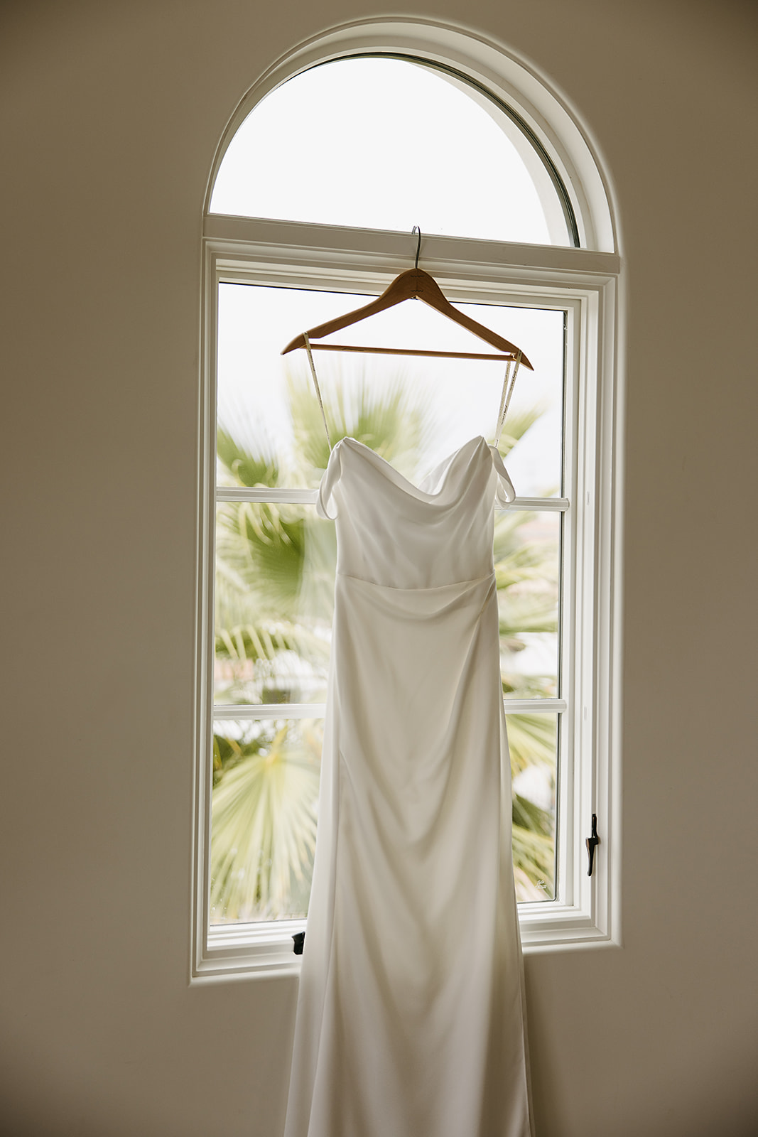 A white wedding dress hangs on a wooden hanger in front of an arched window with a palm tree visible outside.