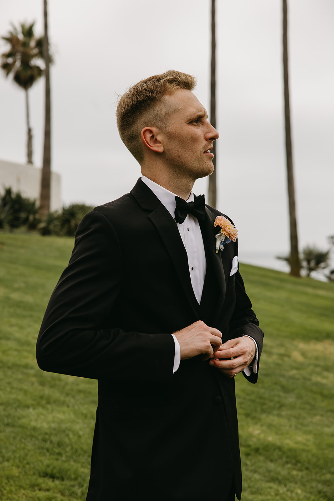 A man in a black tuxedo stands outdoors on grass, adjusting his jacket. There are tall palm trees in the background.