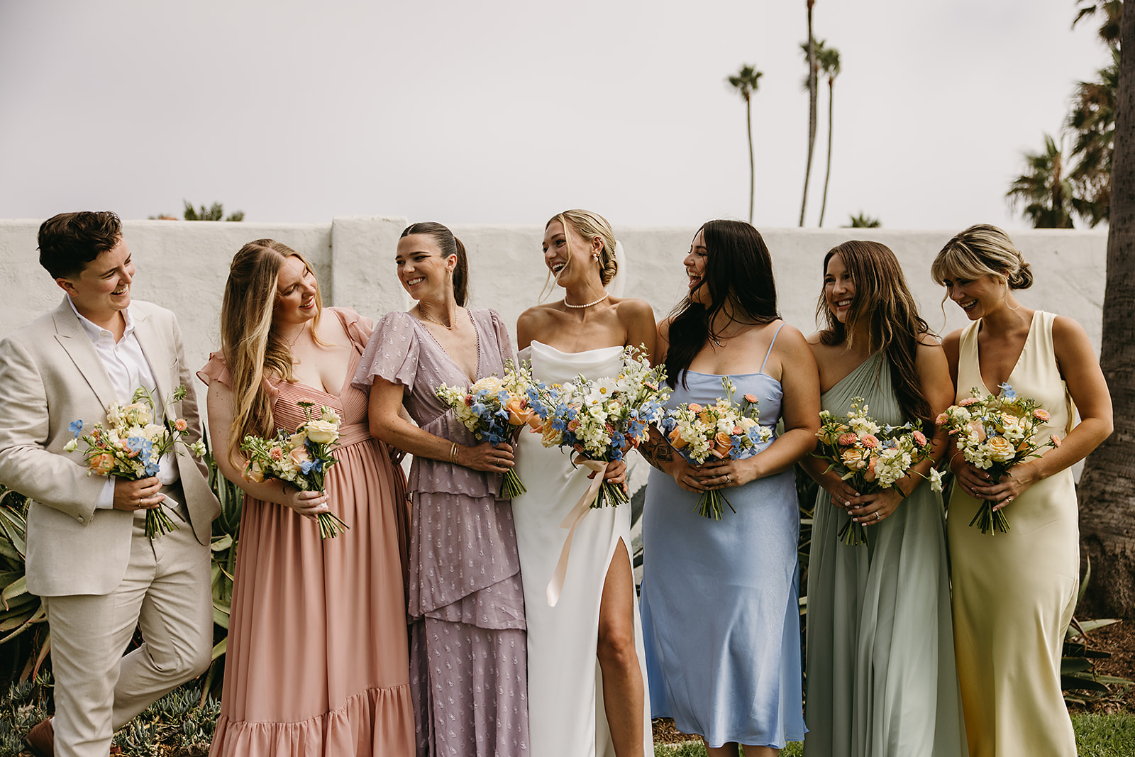  A group of people in formal attire stand together outdoors, smiling and holding bouquets.