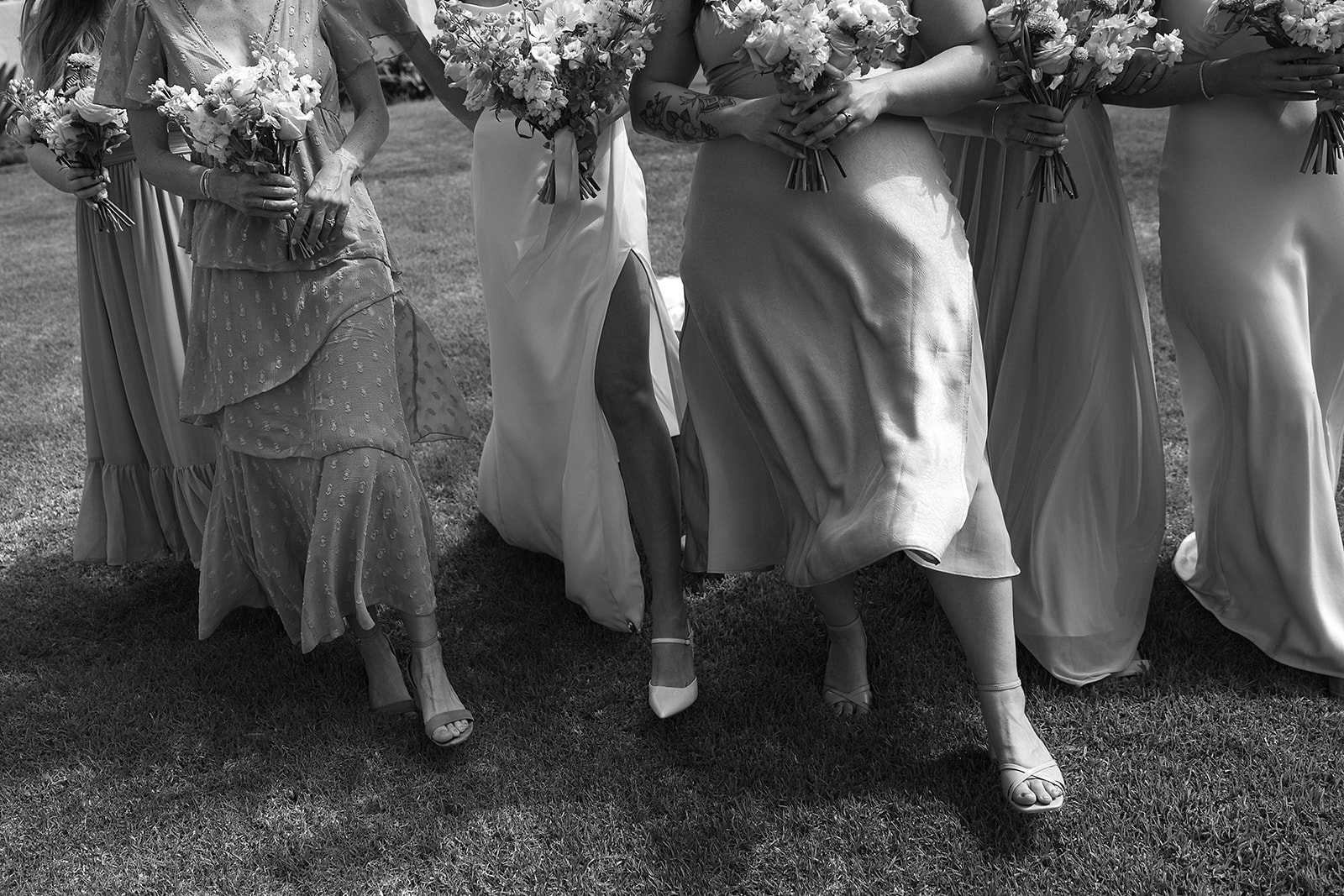 A group of people in formal attire stand together outdoors, smiling and holding bouquets.