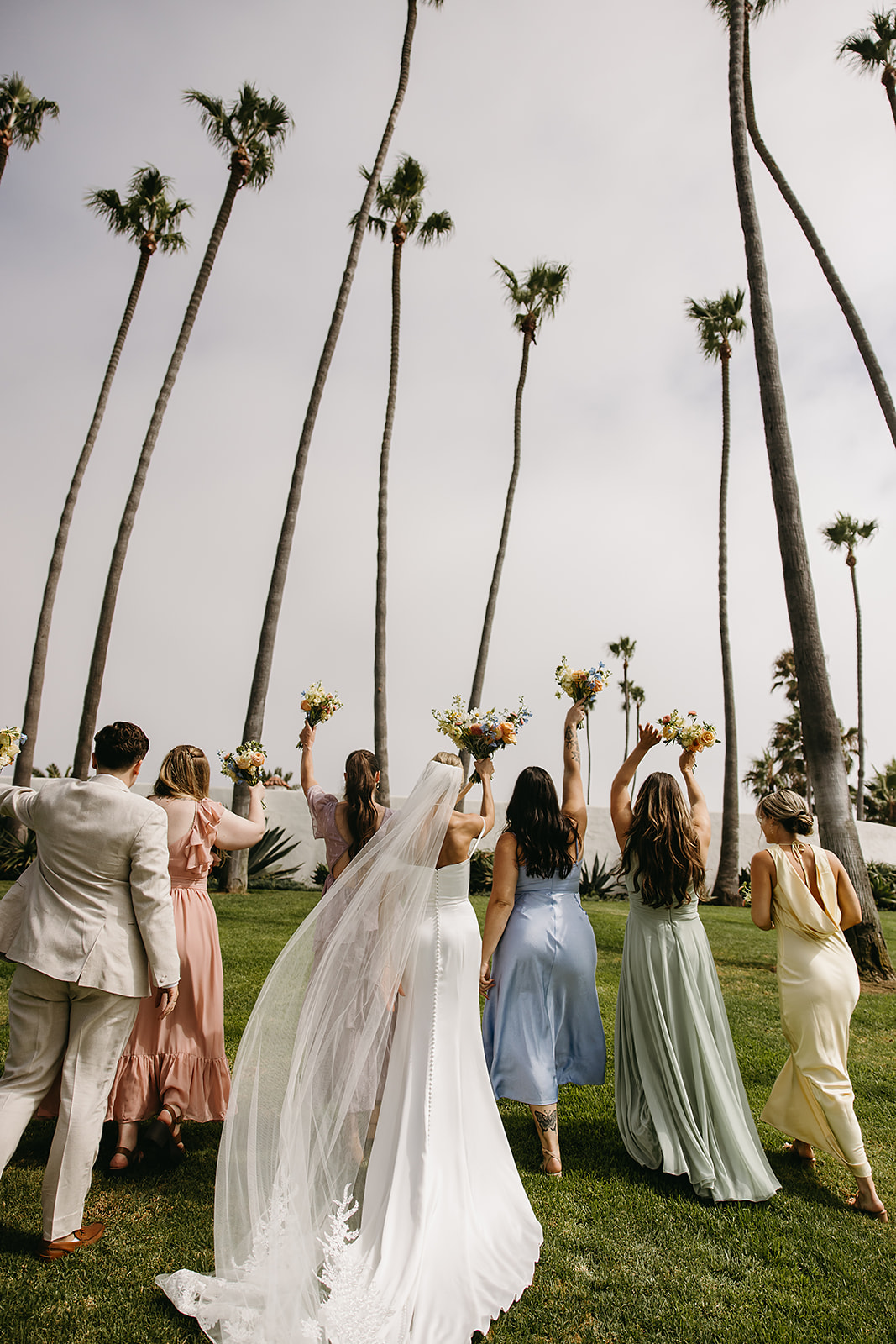 A group of people in formal attire stand together outdoors, smiling and holding bouquets.