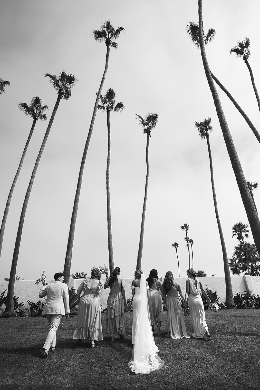 A group of people in formal attire stand together outdoors, smiling and holding bouquets.