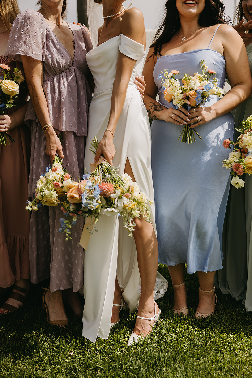 A group of people in formal attire stand together outdoors, smiling and holding bouquets.