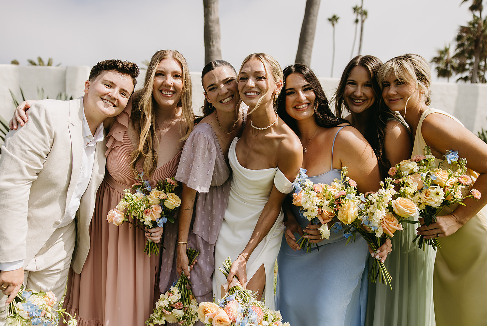 A group of people in formal attire stand together outdoors, smiling and holding bouquets.