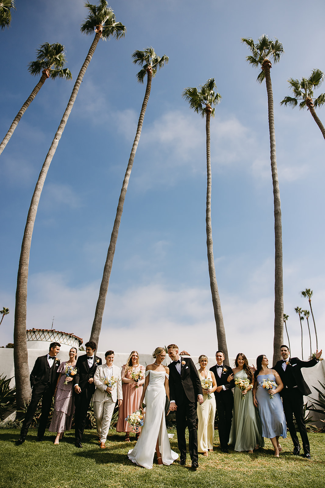 A group of people in formal attire stand together outdoors, smiling and holding bouquets.