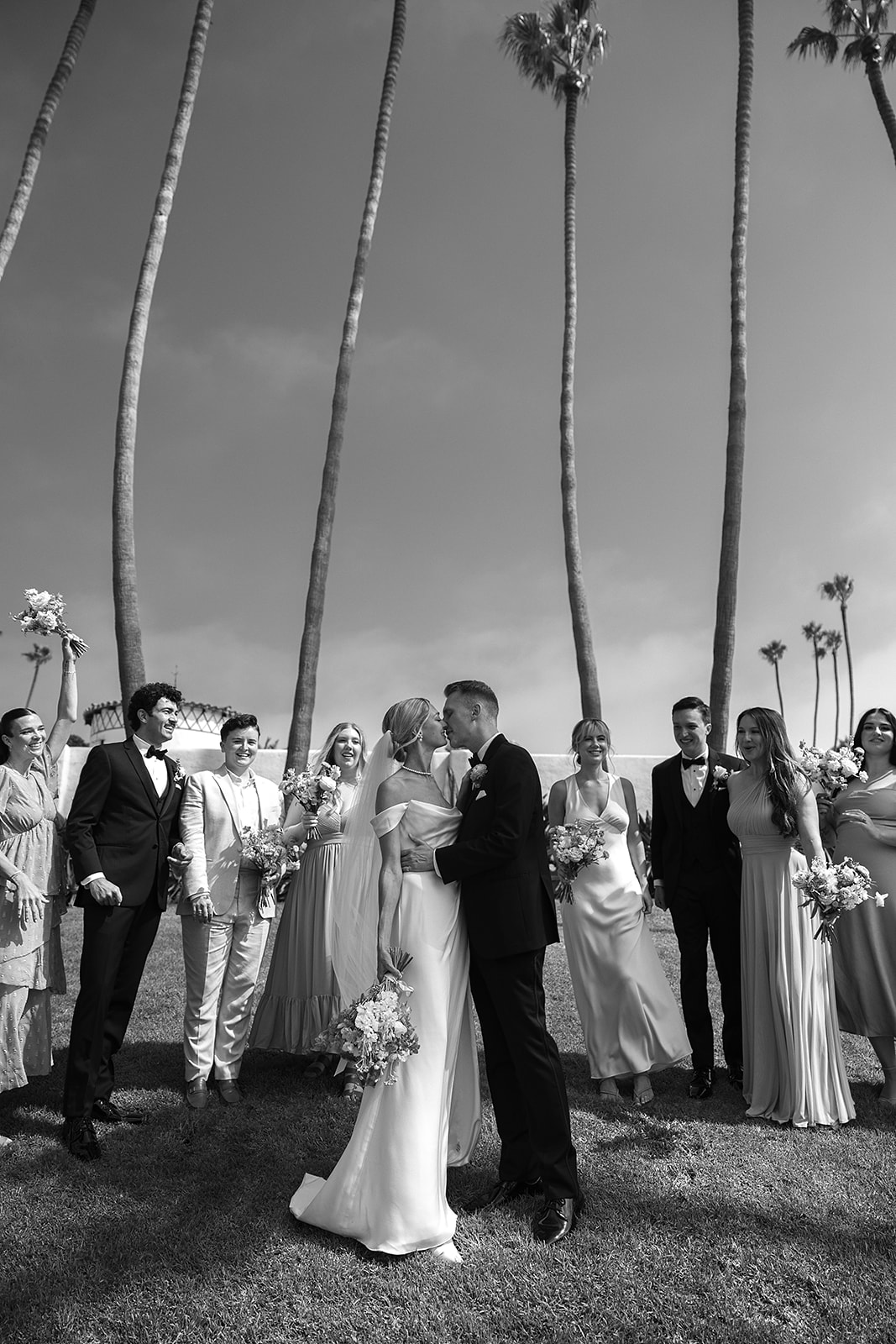 A group of people in formal attire stand together outdoors, smiling and holding bouquets.