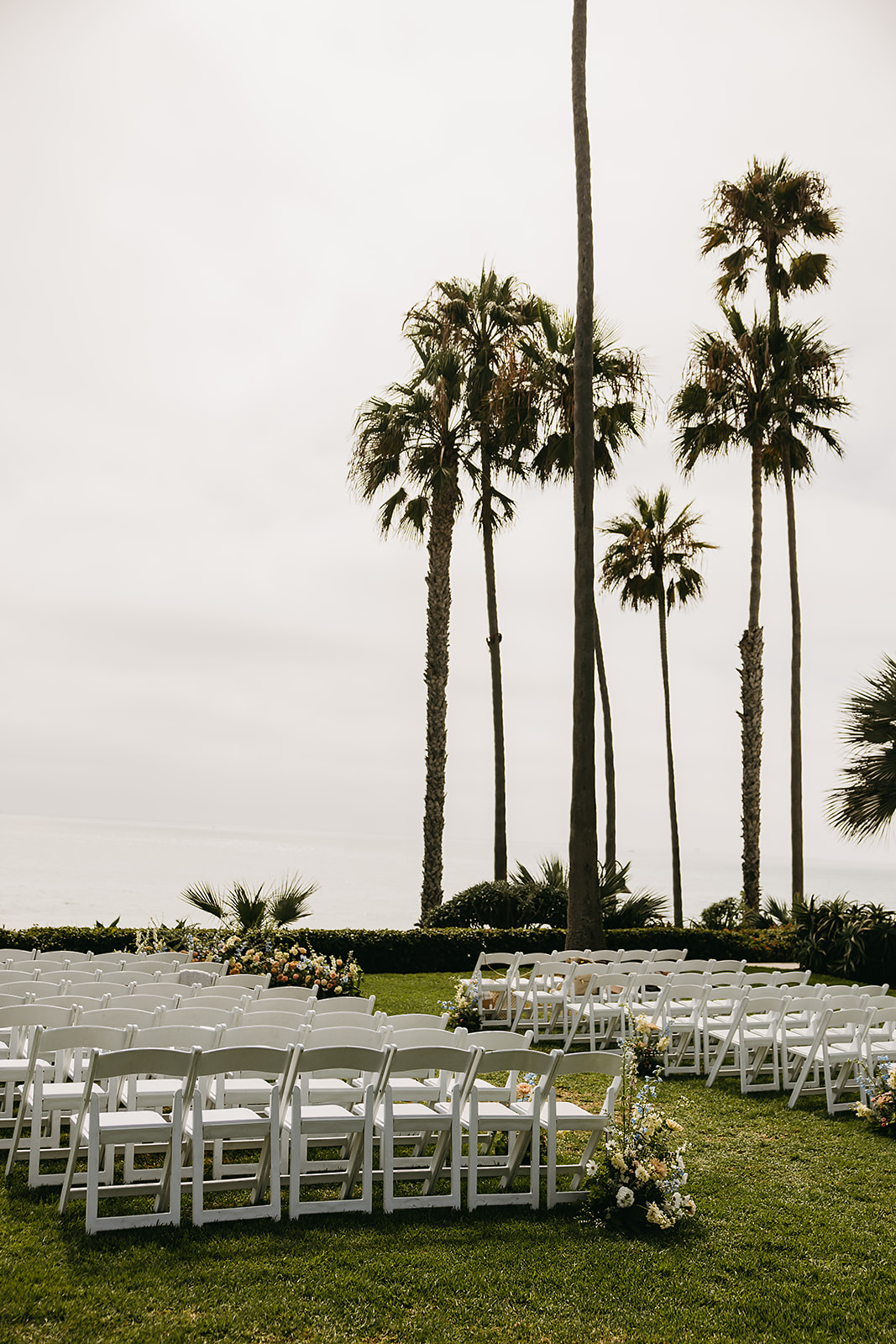 Outdoor wedding ceremony setup with white chairs, floral arrangements, and tall palm trees in the background under a cloudy sky at Ole Hanson