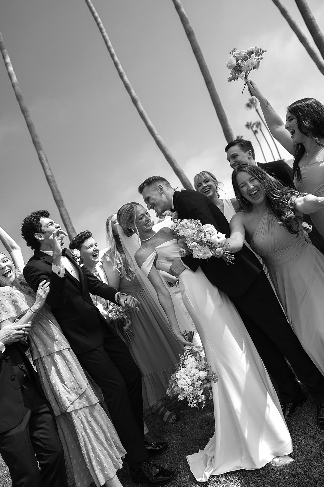 A group of people in formal attire stand together outdoors, smiling and holding bouquets.