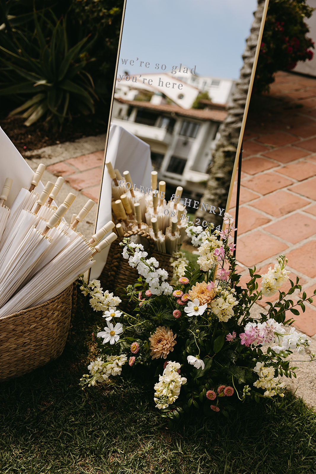 A decorative mirror reflects a building. White umbrellas are displayed in baskets beside a floral arrangement.