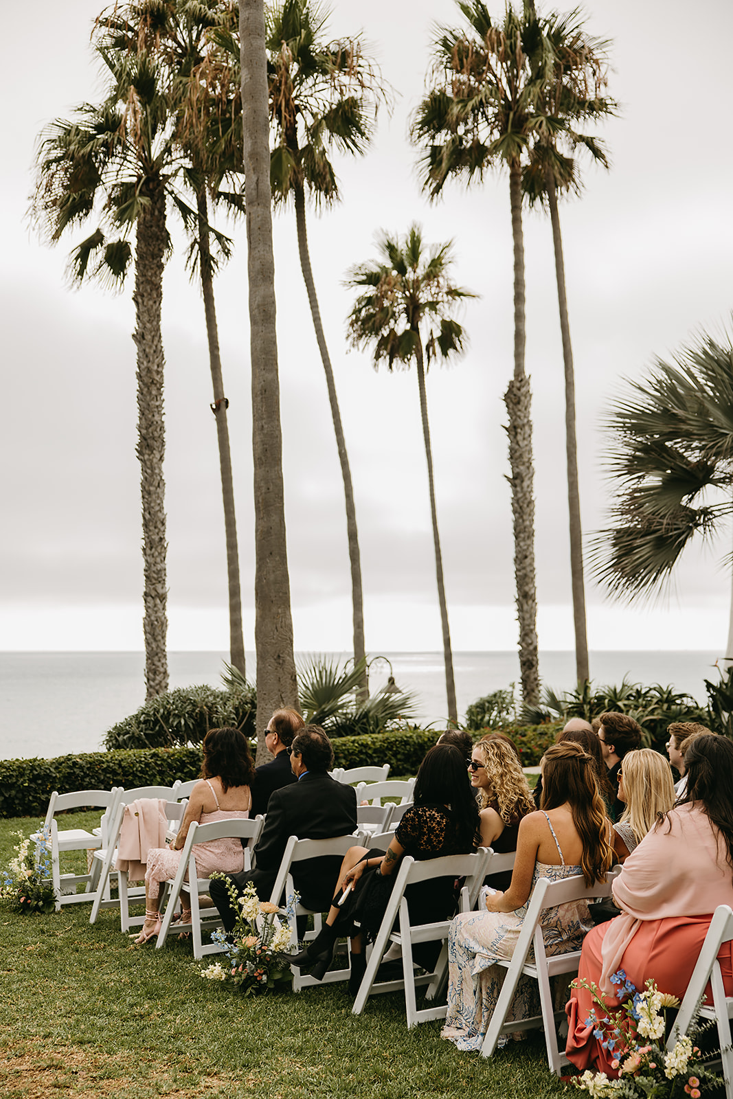 People seated on white chairs outdoors at a wedding ceremony by the sea, with palm trees in the background.