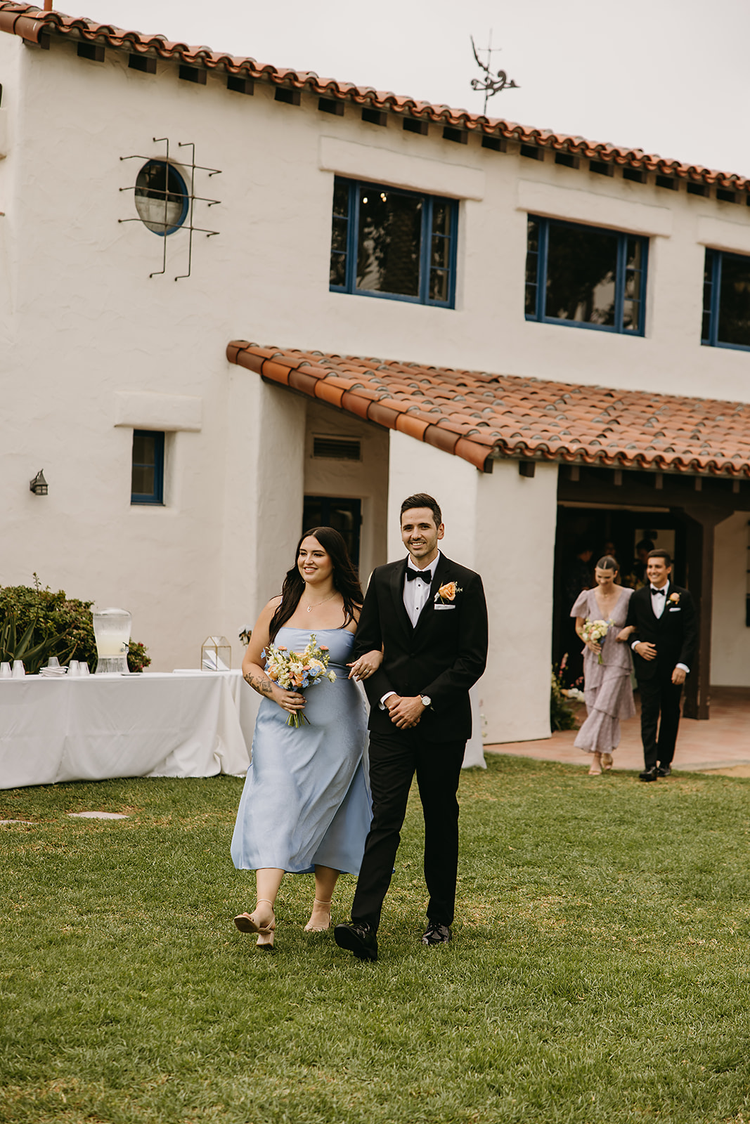 A couple in formal attire walks outdoors on grass near a white building with red-tiled roofs. They seem to be part of a procession, followed by another couple in the background.