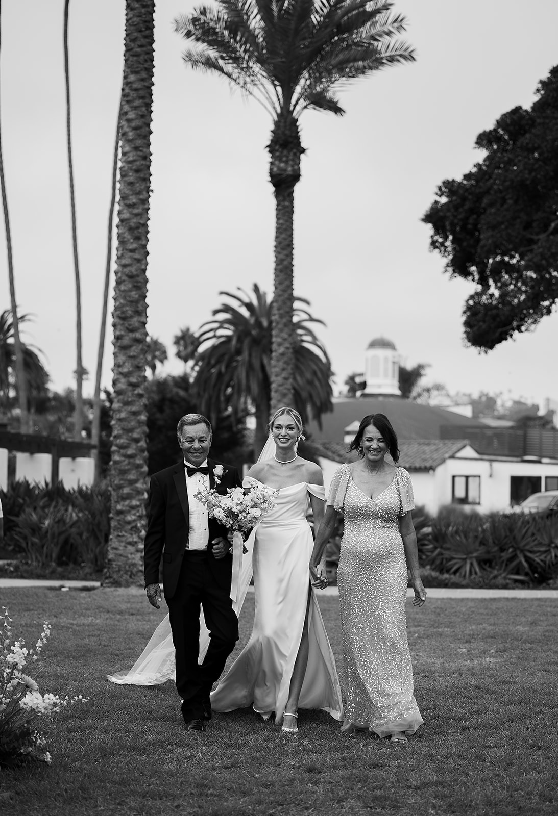 A bride in a wedding dress walks between two people on a lawn with palm trees in the background.