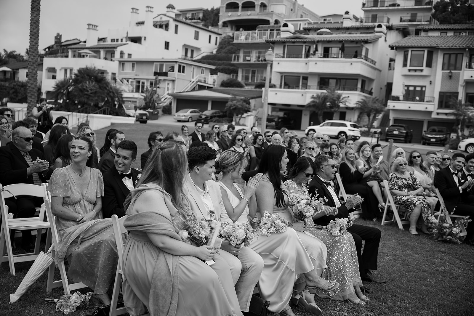 A wedding ceremony audience seated outdoors, with guests dressed formally. Buildings are visible in the background.