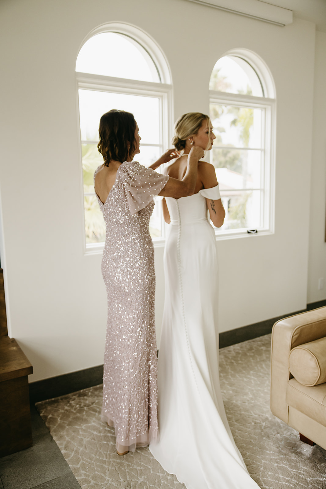 A white wedding dress hangs on a wooden hanger in front of an arched window with a palm tree visible outside.