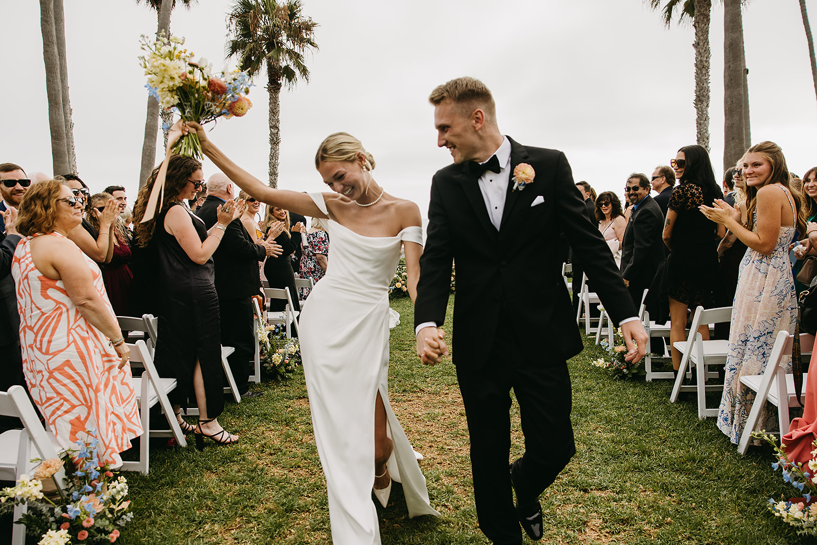 A bride and groom walk down an outdoor aisle, holding hands and smiling, with guests applauding on both sides.