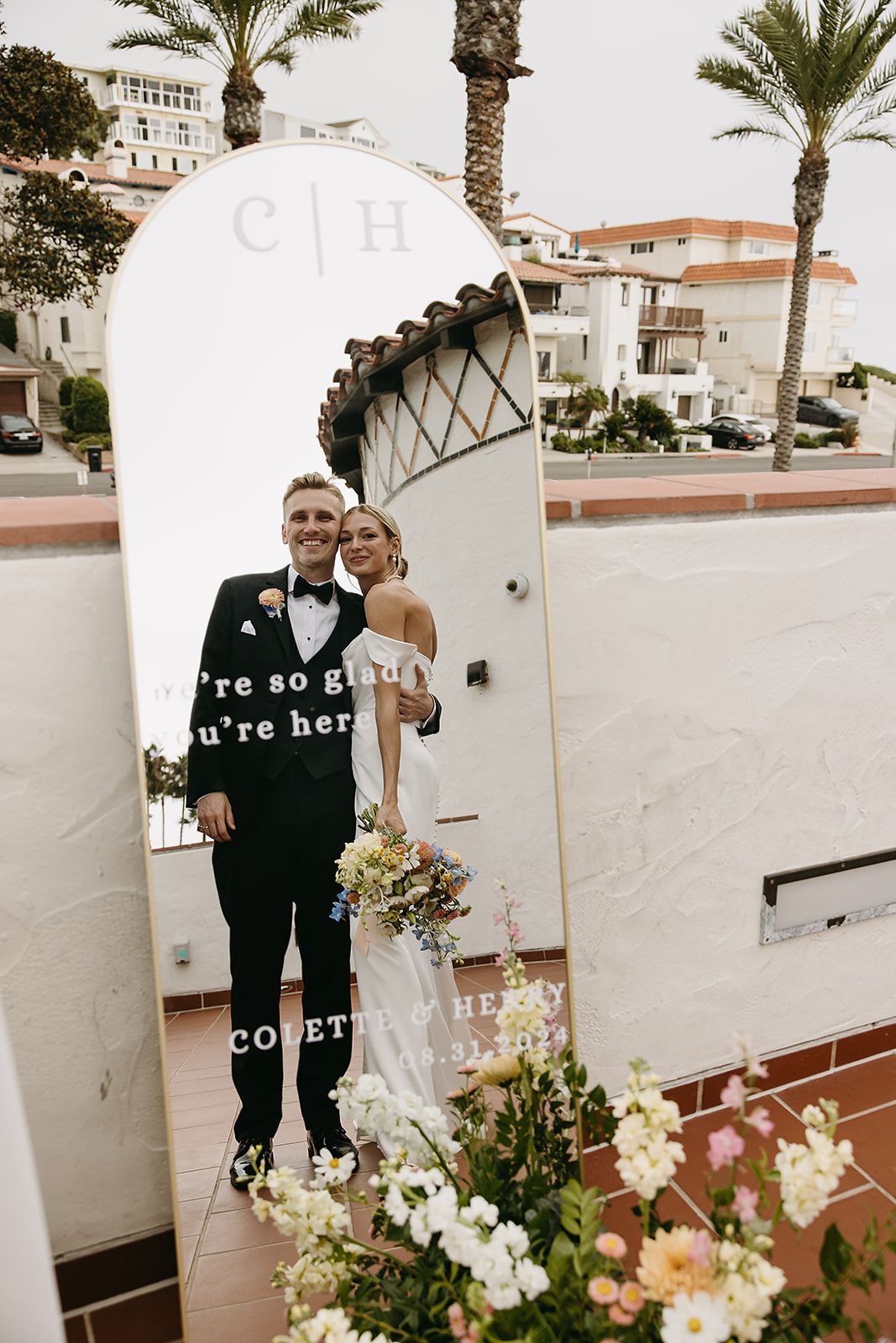 A couple in formal attire poses for a photo, reflected in a tall mirror with flowers at the base. Buildings and palm trees are visible in the background.