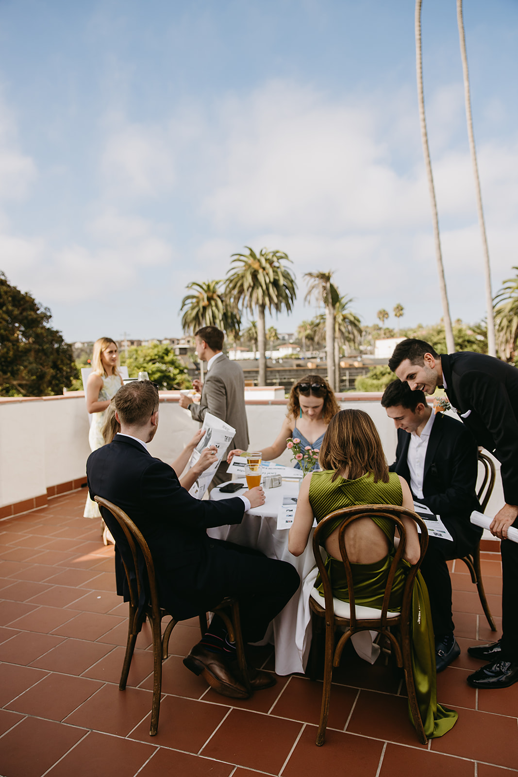 A group of people sitting at an outdoor table on a terrace, with palm trees and blue sky in the background.