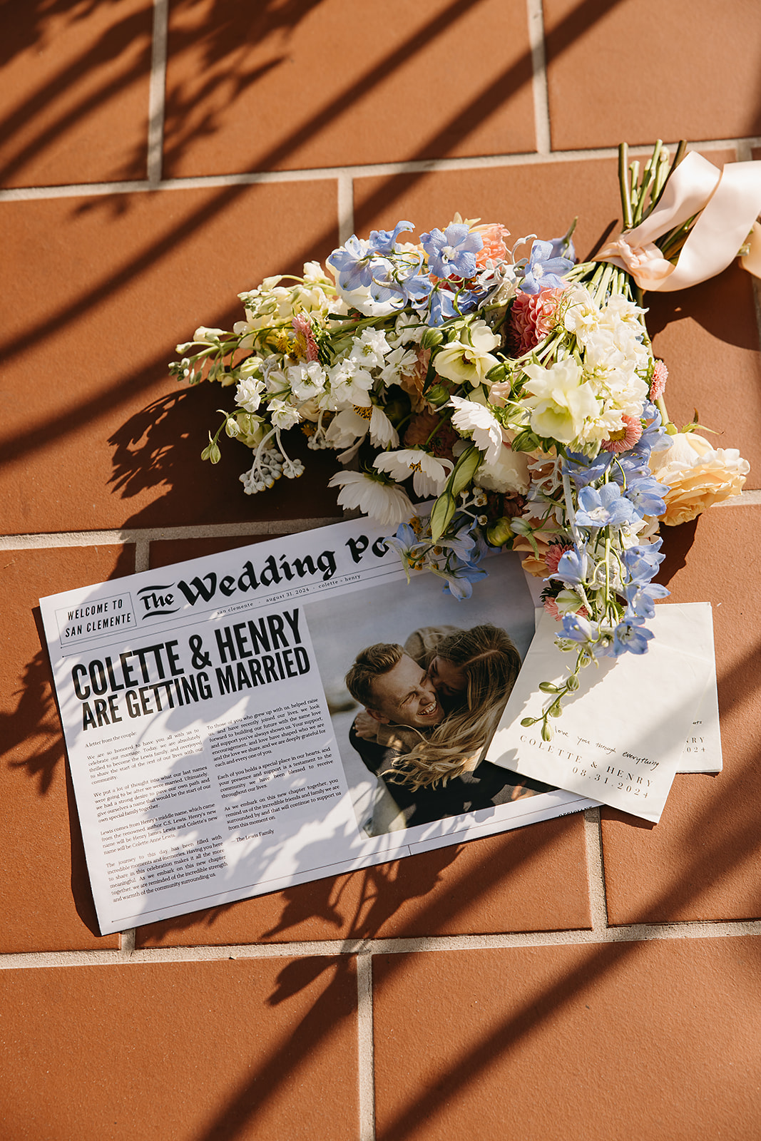 A newspaper titled "The Wedding Post" with a headline about Colette and Henry's wedding lies on a tile floor next to a bouquet of white, blue, and peach flowers.