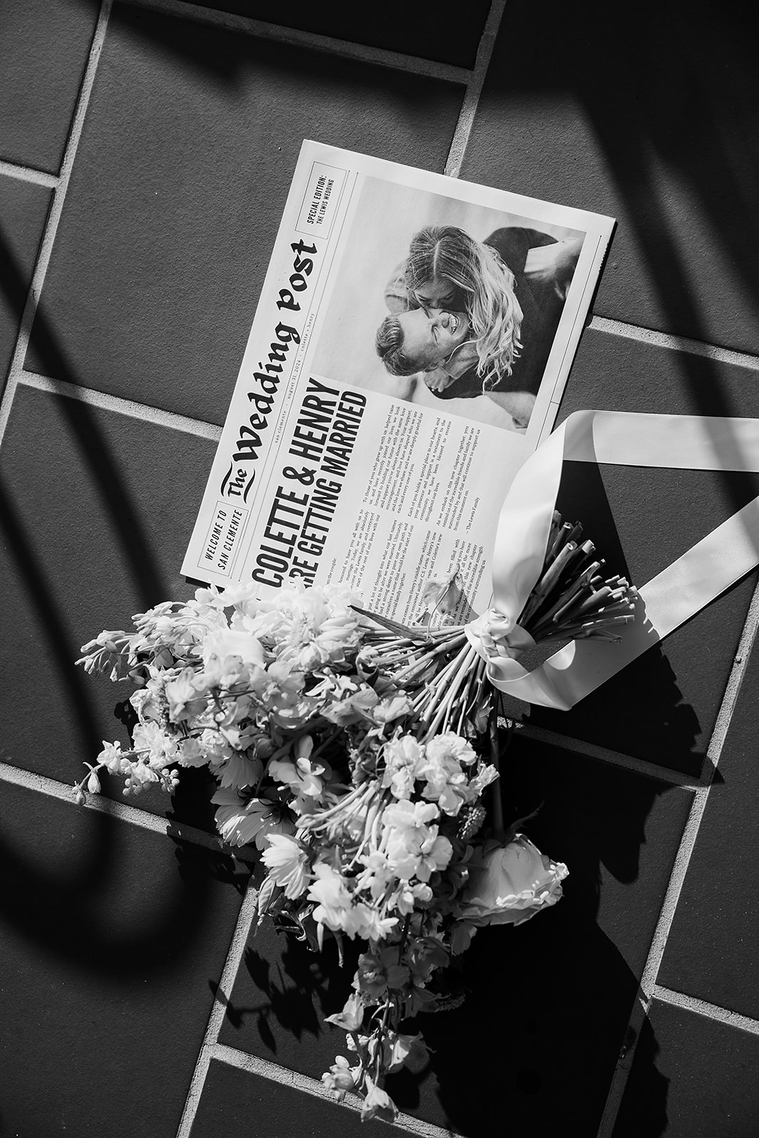 A newspaper titled "The Wedding Post" with a headline about Colette and Henry's wedding lies on a tile floor next to a bouquet of white, blue, and peach flowers.