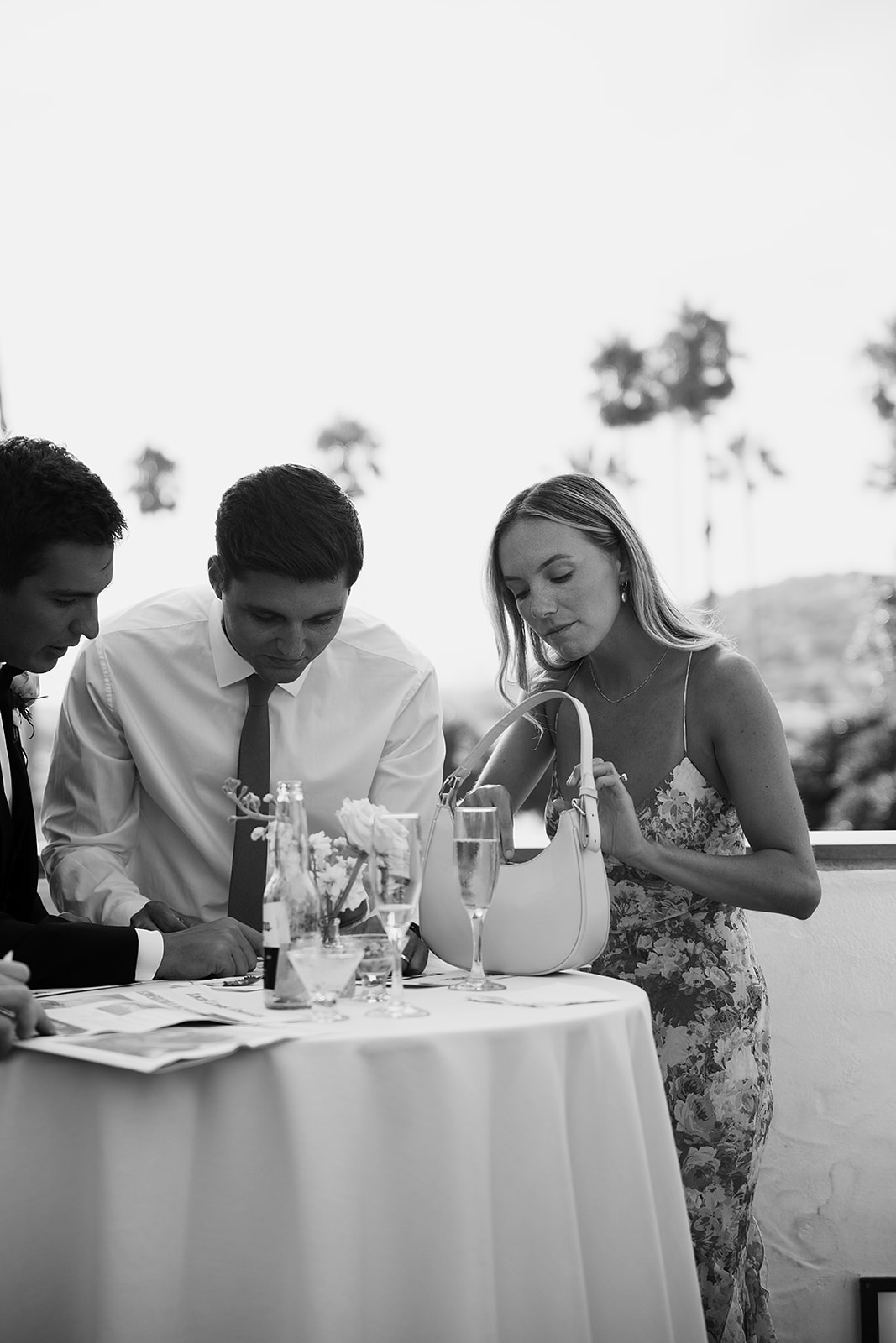 Three people stand around a table with drinks and flowers outdoors. One person is placing something in a handbag.