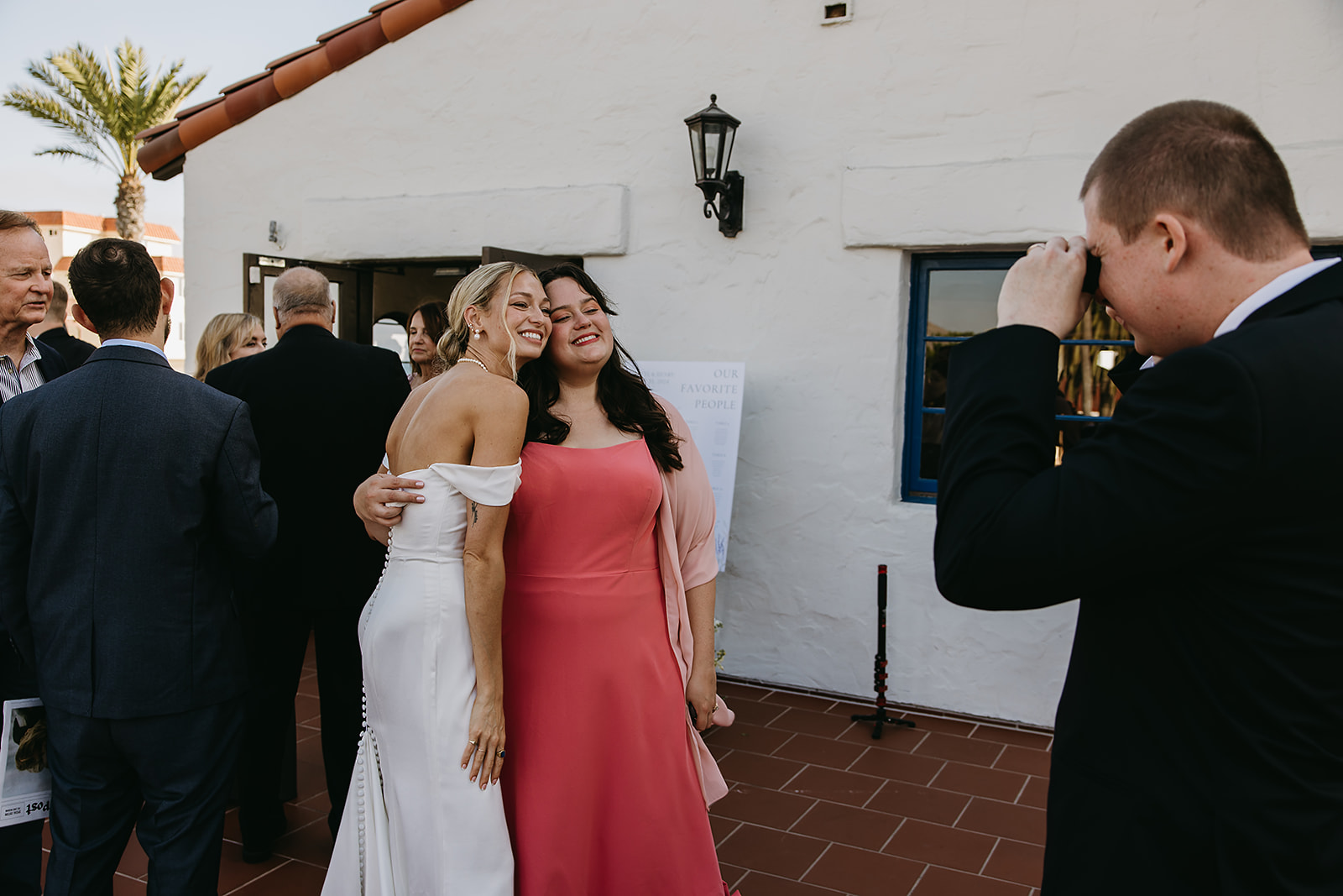 Two women pose for a photo, one in a white dress and the other in a pink dress, while a man takes their picture. People are gathered around a white building with a tile roof at Ole Hanson