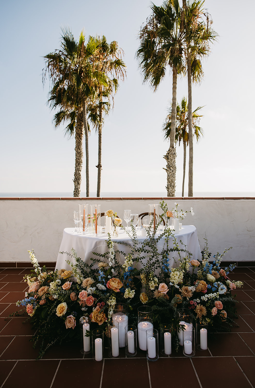 A round table set for dining on an outdoor terrace with palm trees in the background. The foreground features a floral arrangement and candles on the ground at Ole Hanson