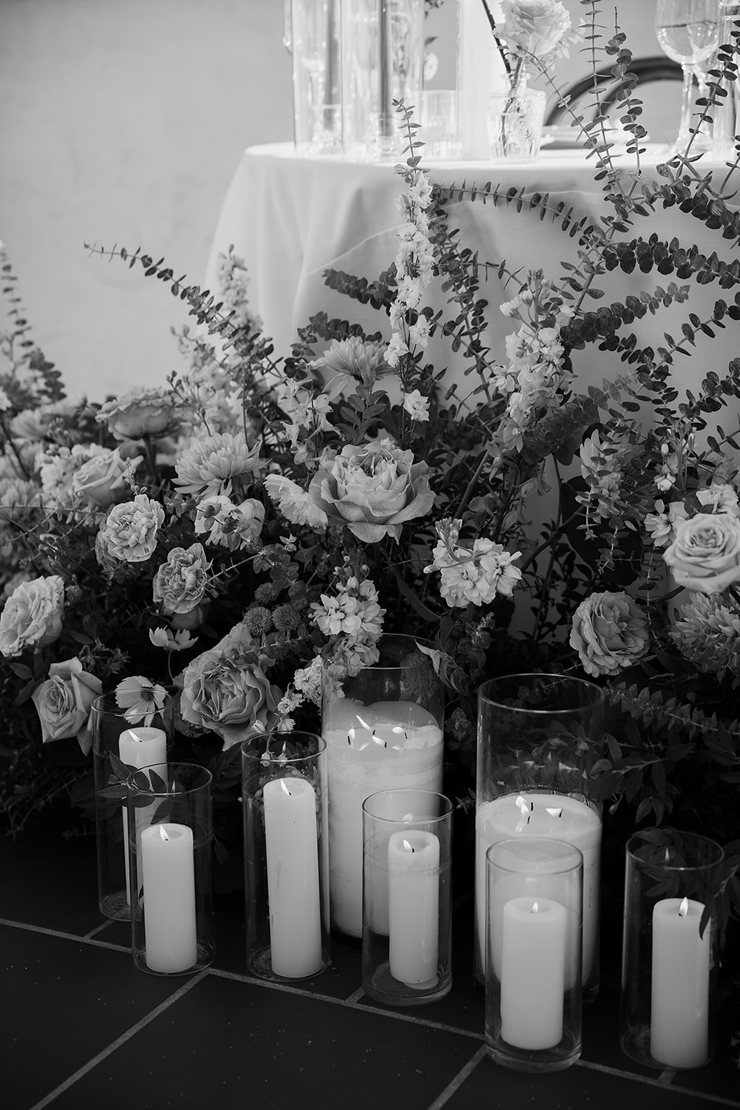 A black and white image of a floral arrangement with roses and ferns, placed in front of tall candles in glass holders.