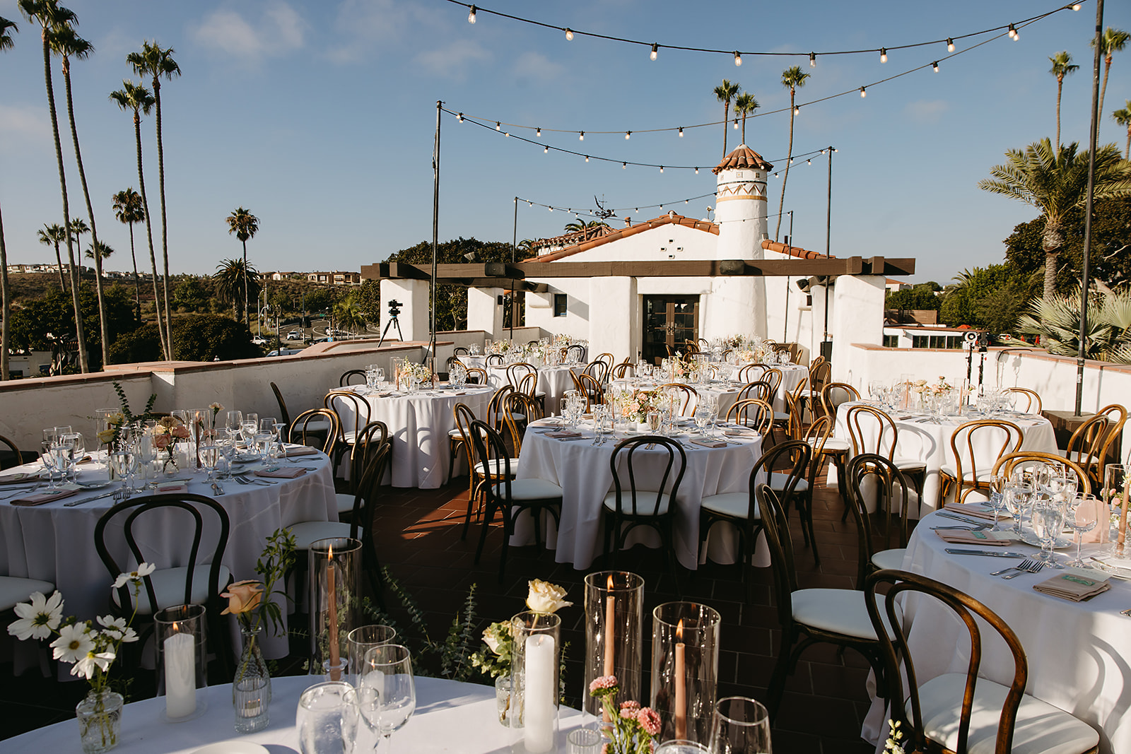 Outdoor wedding reception at Ole Hanson setup with round tables and wooden chairs, decorated with flowers and string lights, set against a sunny sky with palm trees in the background.