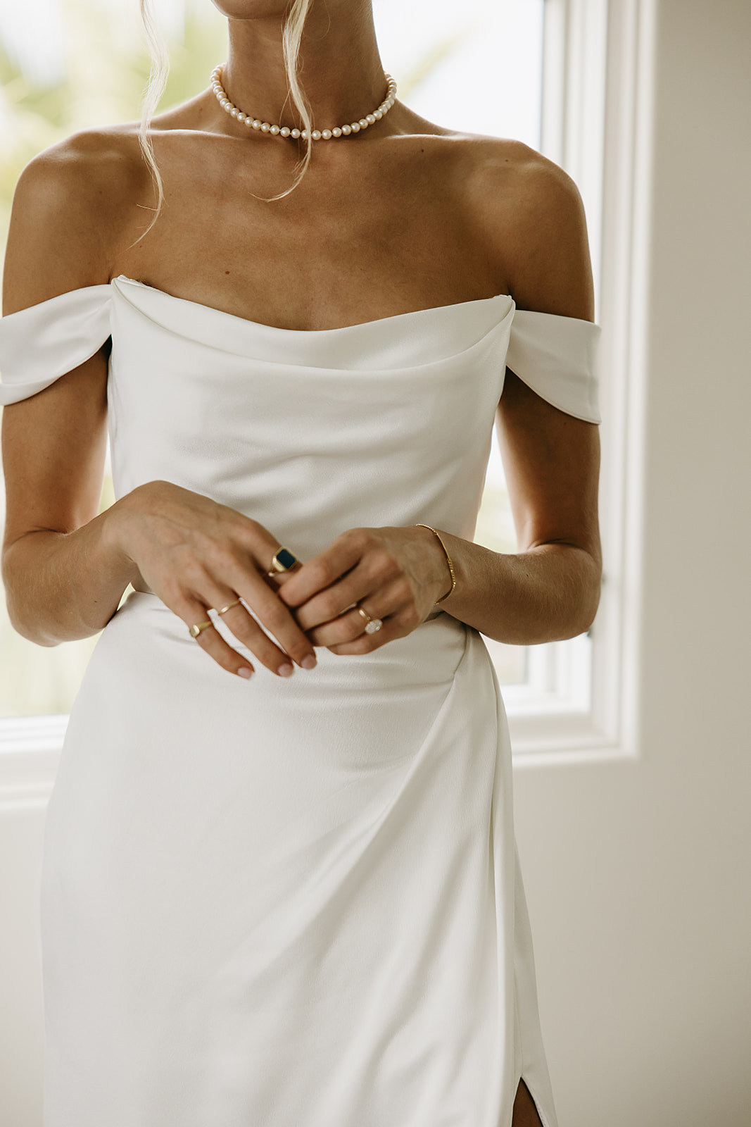 Person in an off-the-shoulder white dress stands by a window, wearing a pearl necklace and a ring.