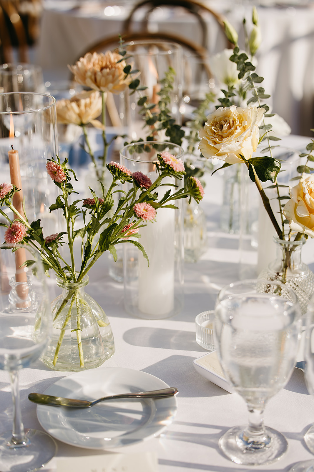 A table set for an event with glass vases holding pink and yellow flowers, glassware, cutlery, and plates on a white tablecloth.