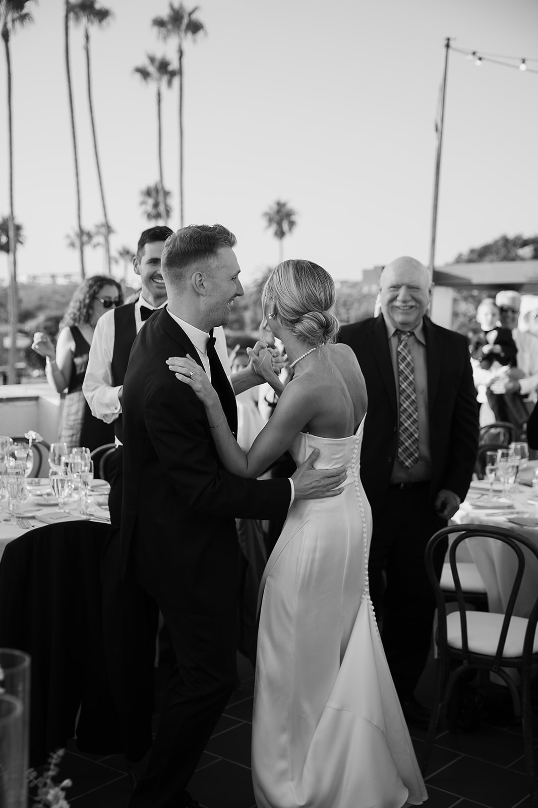 A couple dances closely at an outdoor event with tables and palm trees in the background. Guests watch them.