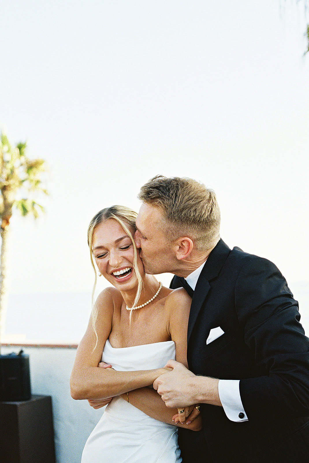 A bride in a white dress and veil smiles at a groom in a black suit as they stand close together on a beach.