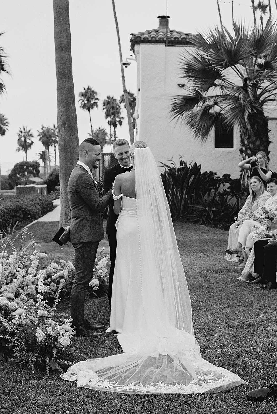 A couple is getting married outdoors, with the bride in a long veil and the groom in a suit, surrounded by flowers and guests seated nearby.