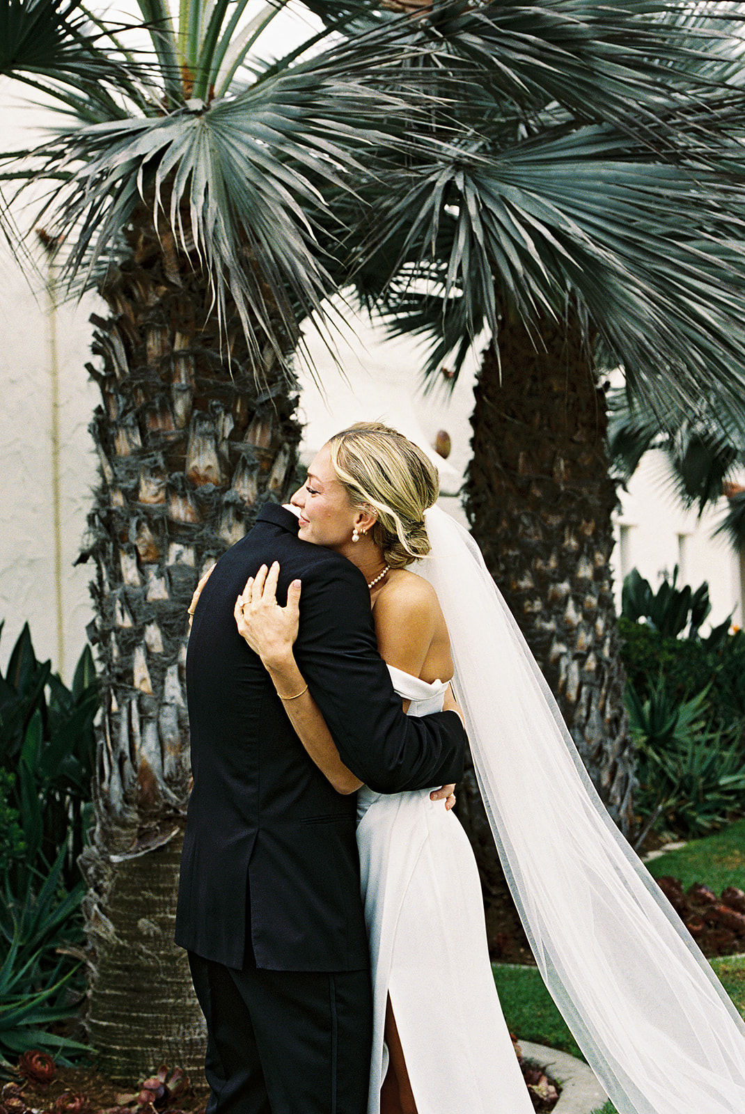 A bride in a white dress and veil smiles at a groom in a black suit as they stand close together on a beach.