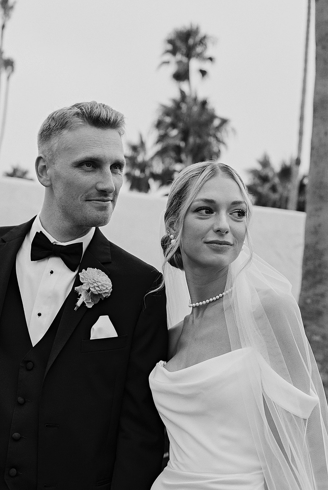 A bride in a white dress and veil smiles at a groom in a black suit as they stand close together on a beach.