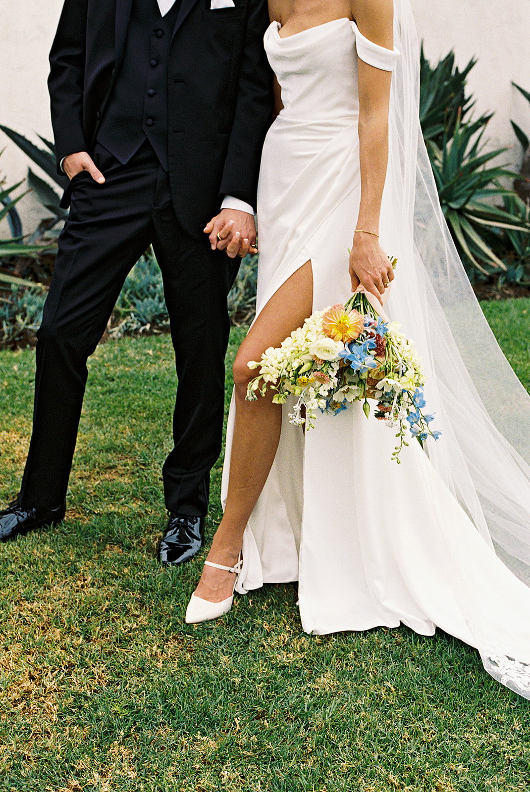 A bride in a white dress and veil smiles at a groom in a black suit as they stand close together on a beach.