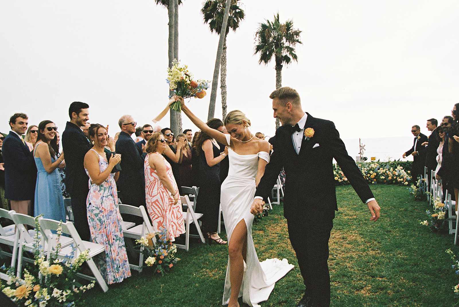 A newlywed couple walks down the aisle outdoors, surrounded by guests. The bride holds a bouquet, and palm trees are in the background.