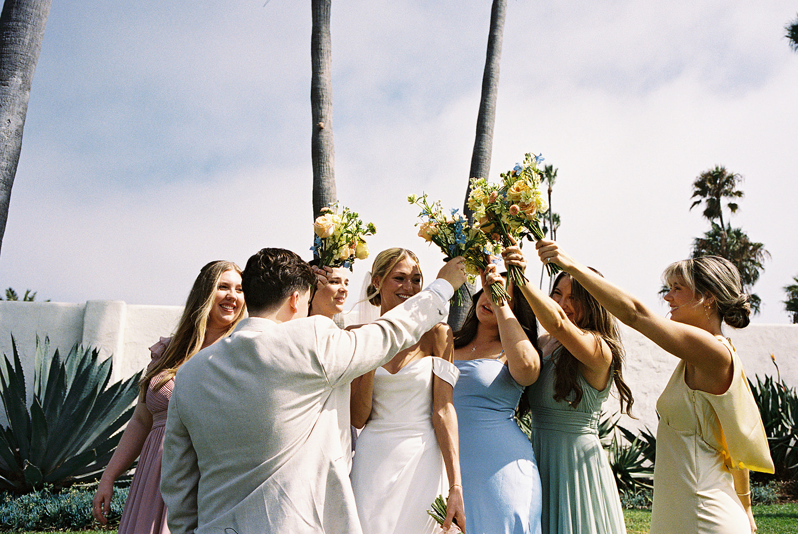 A group of people in formal attire raises flowers outdoors. Palm trees and a concrete wall are in the background.