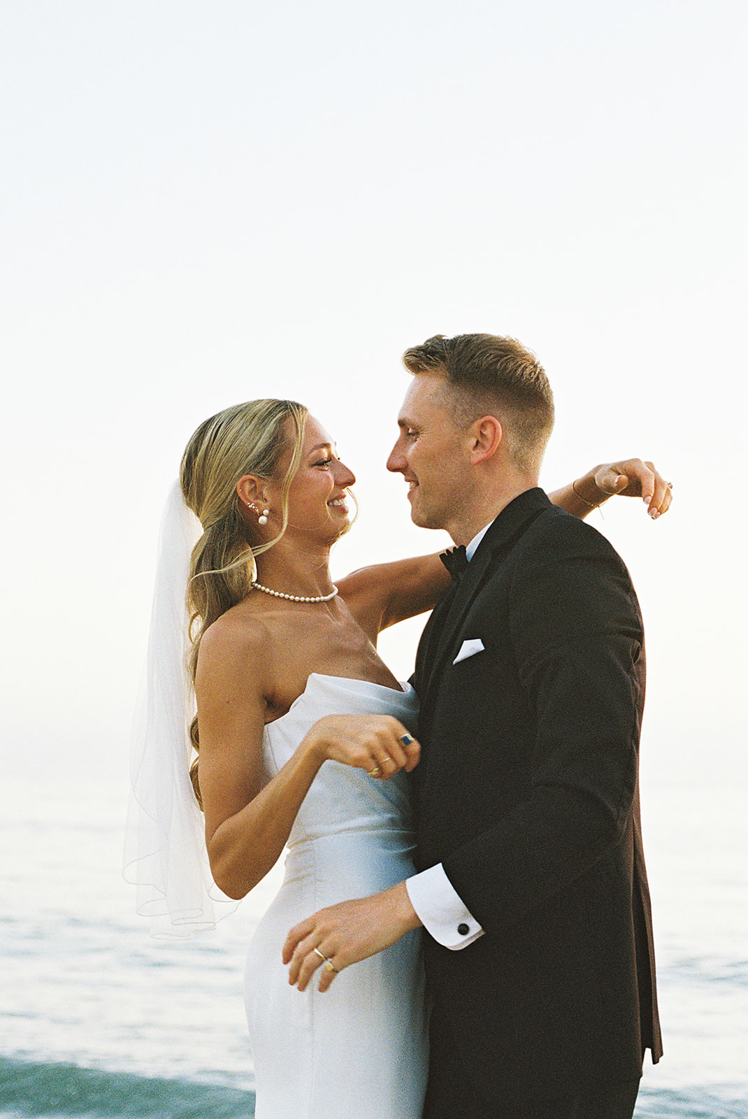 A bride in a white dress and veil smiles at a groom in a black suit as they stand close together on a beach.