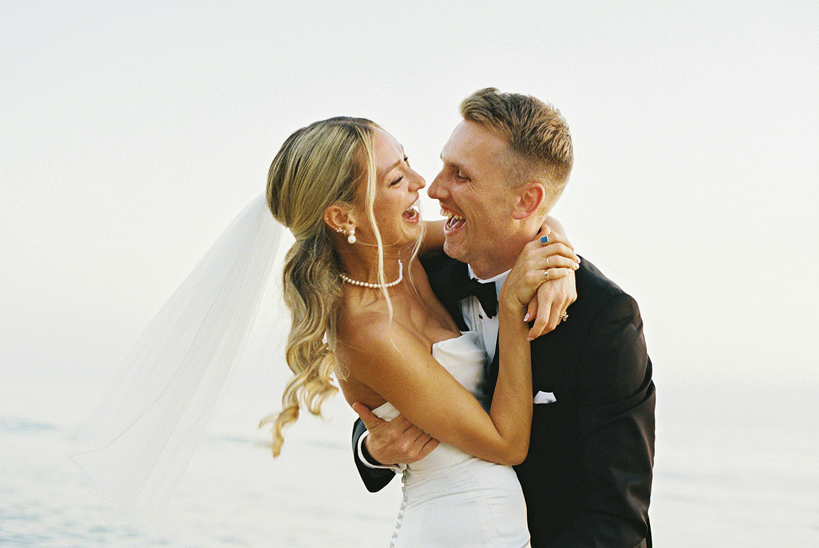 A bride and groom embrace and smile at each other against a soft, blurred background.