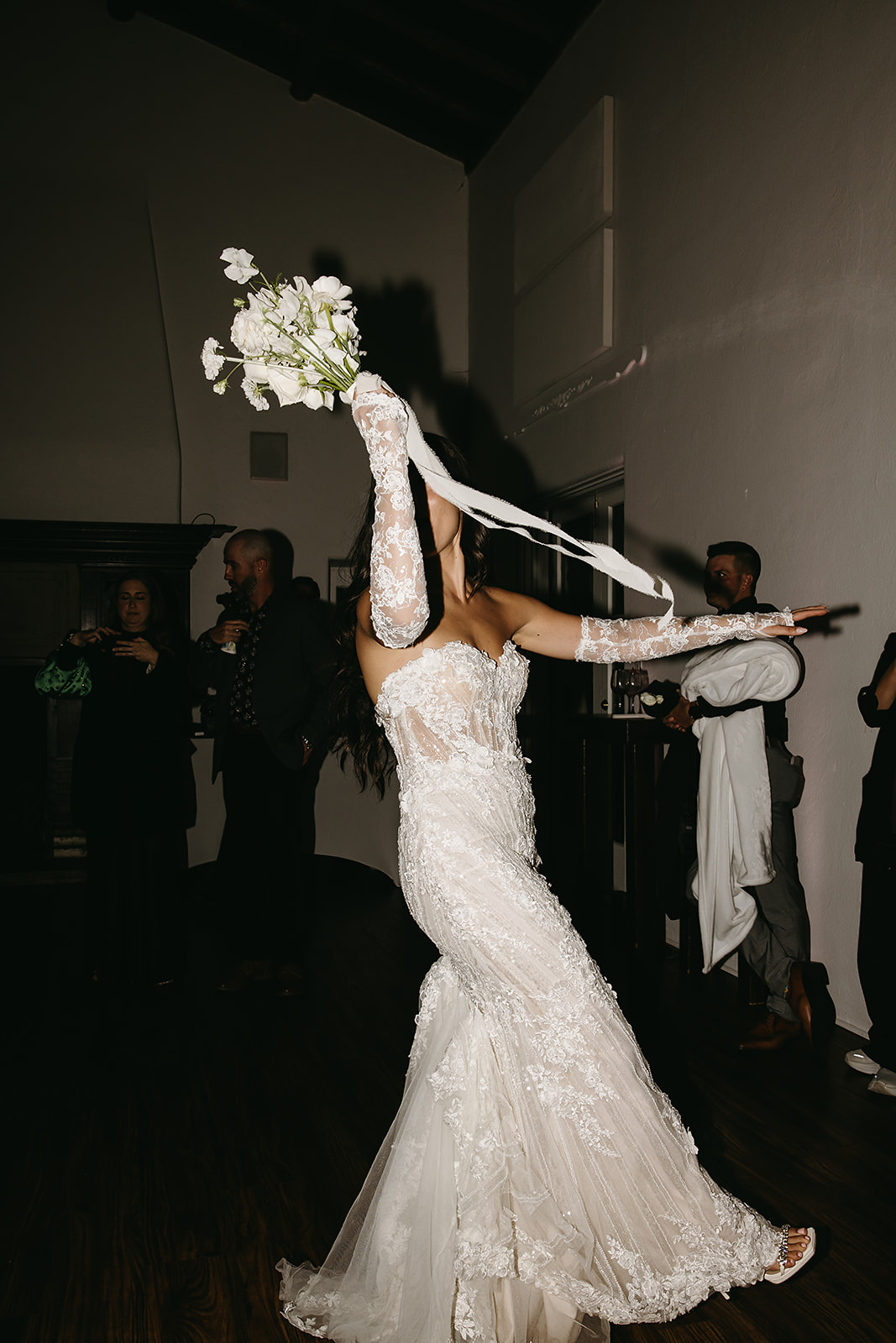 Bride in a white lace dress tosses her bouquet over her shoulder in a dimly lit room as guests look on.