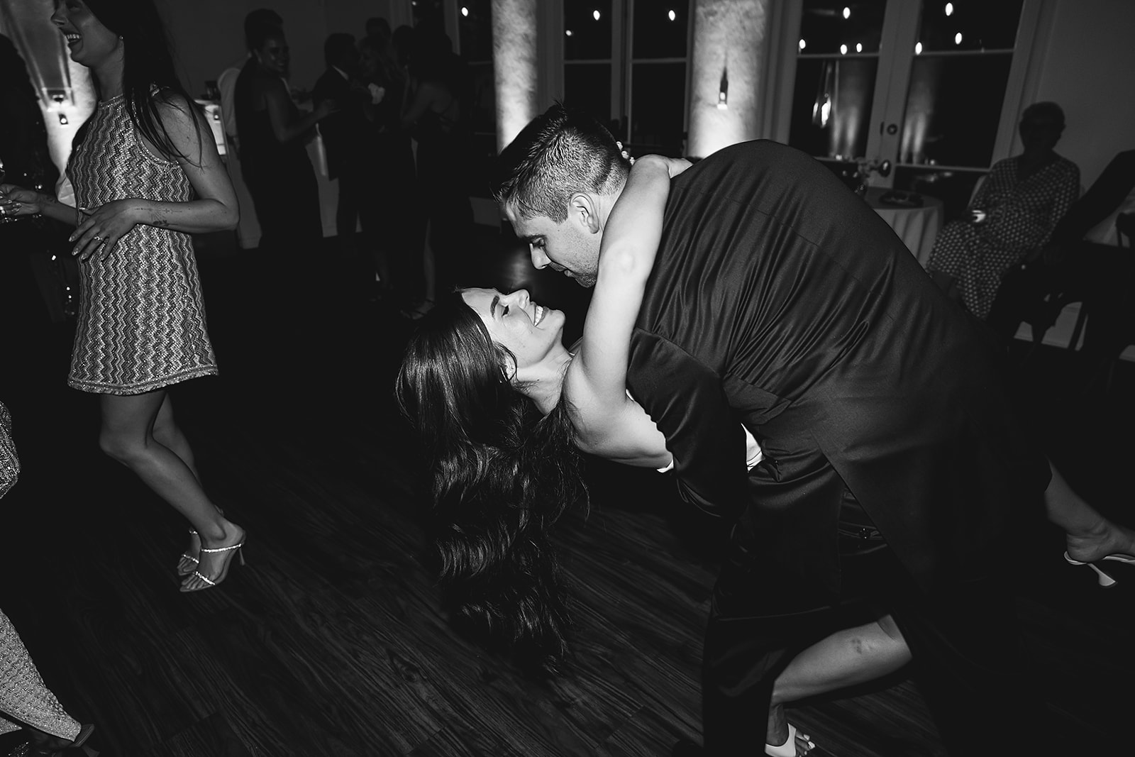 A couple dances closely on a dimly lit dance floor, with the man dipping the woman while others dance in the background.