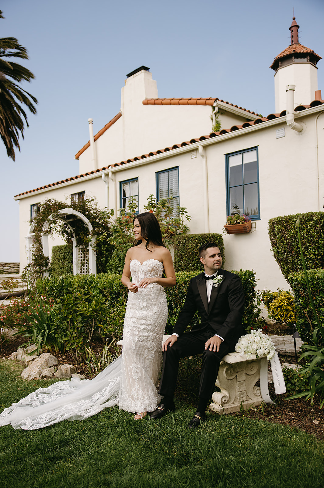 A bride and groom pose outdoors in front of a white house with red roof tiles. The bride stands holding a bouquet, while the groom sits holding a flower at the La Venta Inn