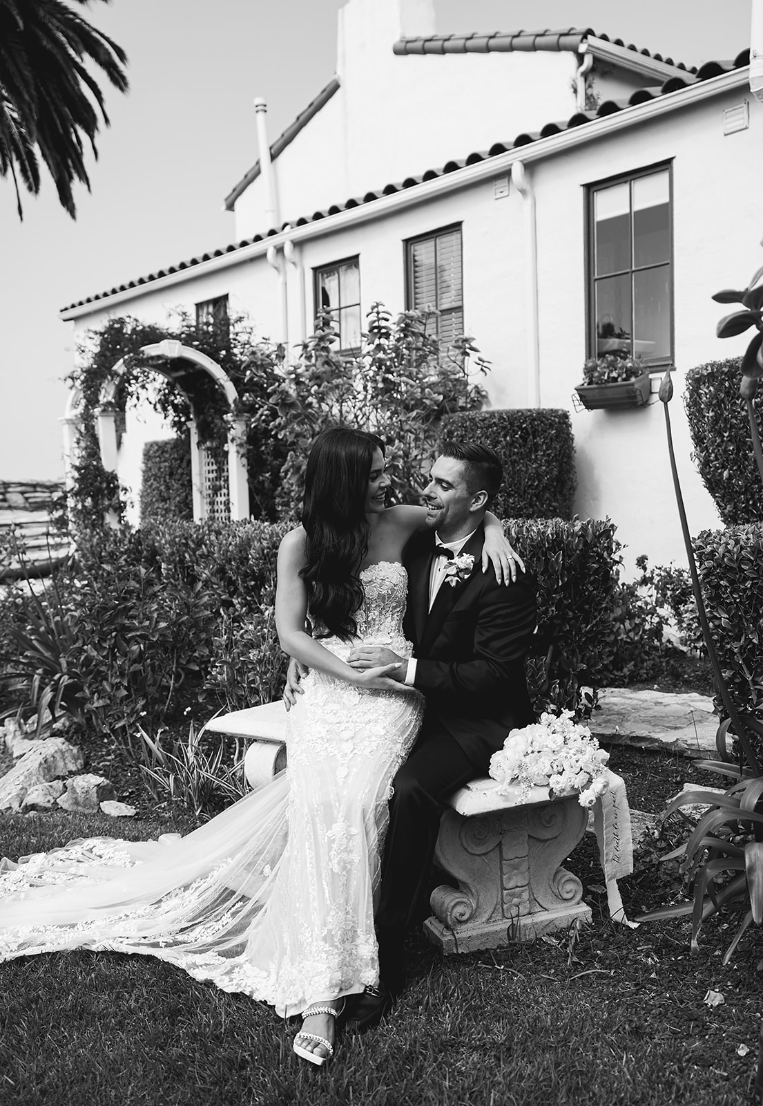 A bride and groom pose outdoors in front of a white house with red roof tiles. The bride stands holding a bouquet, while the groom sits holding a flower at the La Venta Inn