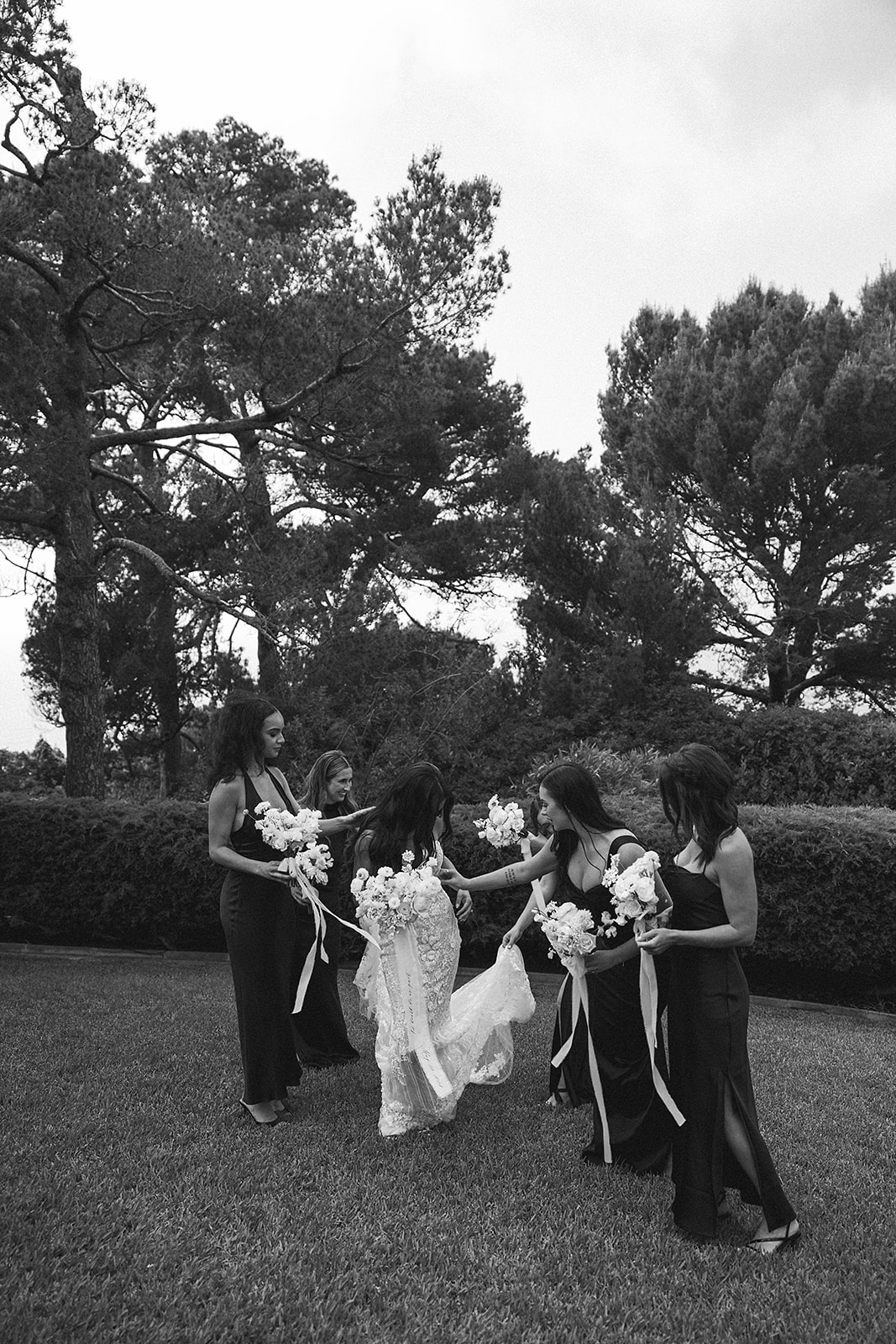 A bride stands with five bridesmaids, each holding white flower bouquets. The women are dressed in black dresses, and they are outdoors with trees and greenery in the background.