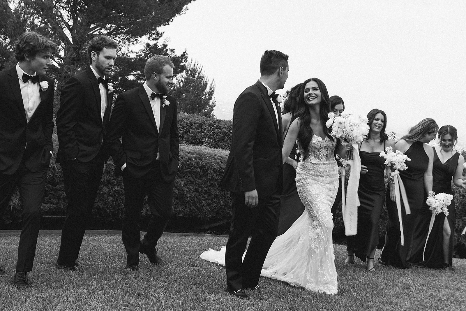 A bride and groom walk outdoors with their wedding party, who are dressed in black. They are holding white flowers, surrounded by lush greenery and trees.