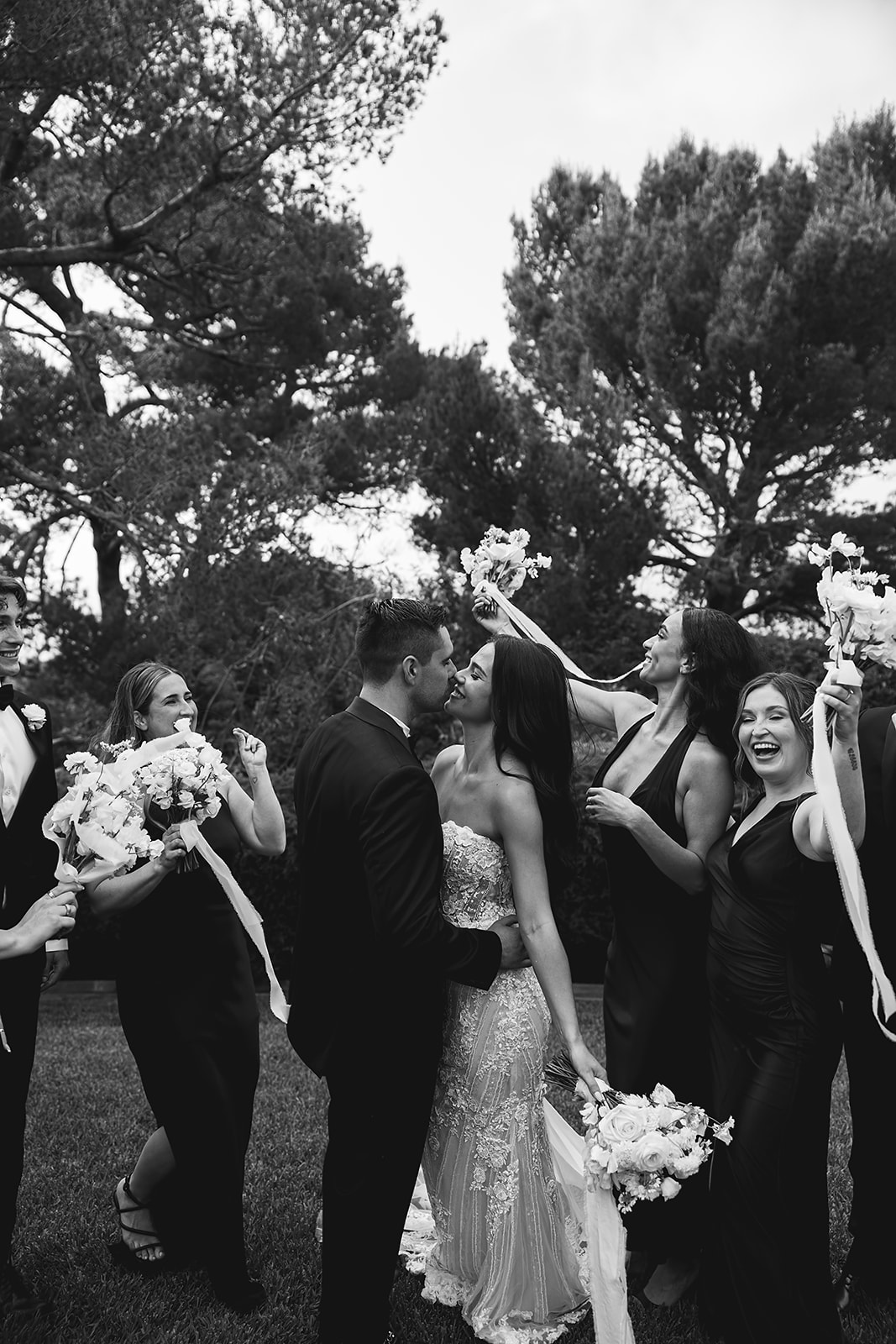 A bride and groom walk outdoors with their wedding party, who are dressed in black. They are holding white flowers, surrounded by lush greenery and trees.